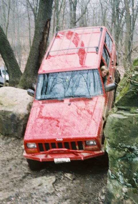 1995 Ford Bronco - This red Jeep Cherokee is navigating a difficult off-road trail, wedged between large rocks and trees.