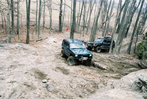 1995 Ford Bronco - Two SUVs tackle a challenging muddy trail through a dense forest during an off-road excursion.