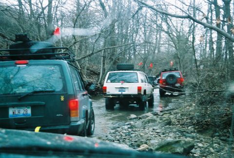 1995 Ford Bronco - Multiple Jeep Cherokees navigate a muddy, wooded off-road trail with red flags marking the path.