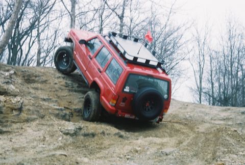 1995 Ford Bronco - Red Jeep Cherokee navigates a challenging off-road hill climb with a spare tire mounted on the rear.