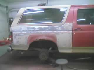 1989 Ford Bronco - Side view of a red two-door SUV with a white removable hardtop, up on jack stands in a garage with the rear wheel removed and the bare brak…