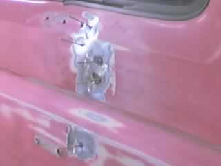 1989 Ford Bronco - Close-up of a faded pink-red door panel during bodywork, where the door handle and lock area has been filled with gray body filler and sand…