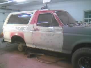 1989 Ford Bronco - Passenger-side view of a two-door Ford Bronco in a garage with the body stripped to gray primer and patchy red paint, the front fender and…