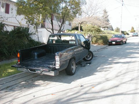 2000 Ford Ranger - A dark gray pickup truck with significant front-end damage is parked on the side of a street, with another car visible in the background.