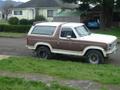 1985 Ford Bronco - A two-tone Ford Bronco is parked on a grassy area next to a house.