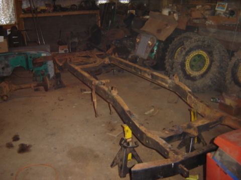 1992 Ford Bronco - A heavily rusted vehicle frame is supported by jack stands in a cluttered garage, with other vehicle parts scattered around.