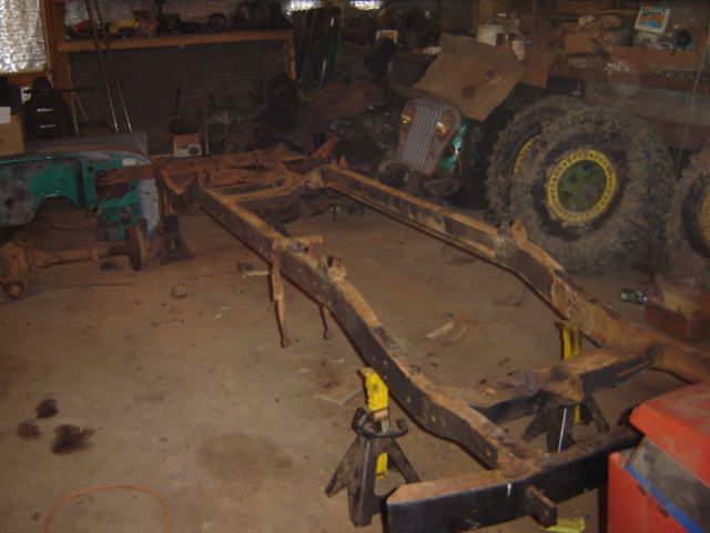 Rusty vehicle frame on jack stands in a garage, with a green fender and large tires visible