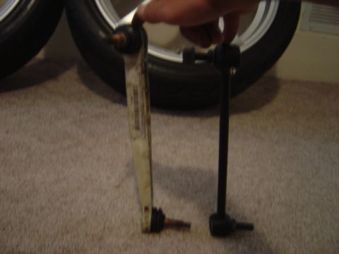 1998 Ford Taurus - Side-by-side comparison of two sway bar end links held upright on carpet: a worn white-painted flat-style link with exposed ball studs next…