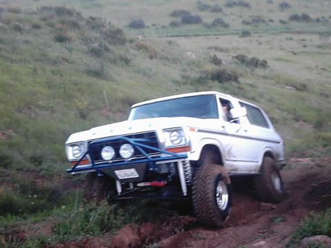 1978 Ford Bronco - A white 1978 Ford Bronco navigates a steep, muddy incline with a custom blue front bumper and large off-road tires.