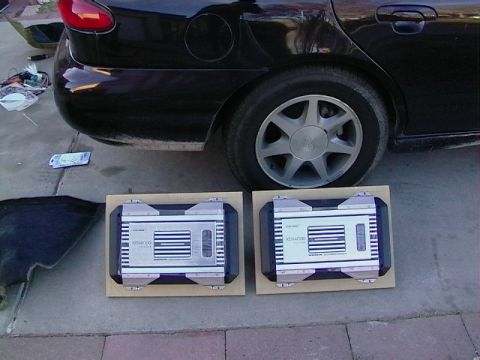 1995 Ford Contour - Two Kenwood car-audio amplifiers sitting in cardboard trays on a driveway in front of a dark sedan with seven-spoke alloy wheels.