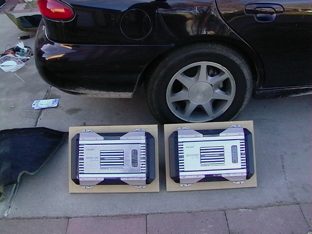 Two Kenwood car amplifiers on a driveway in front of a dark sedan