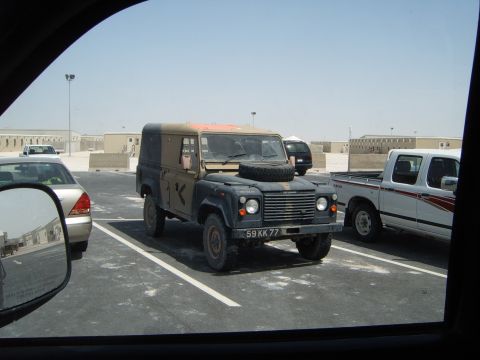 1988 Ford Bronco - A utilitarian Land Rover Defender is parked in a lot, alongside other vehicles under a clear sky.