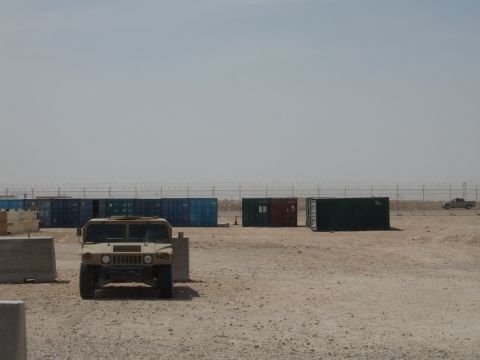 1988 Ford Bronco - A tan Humvee is parked in a lot with shipping containers in the background under a clear sky.