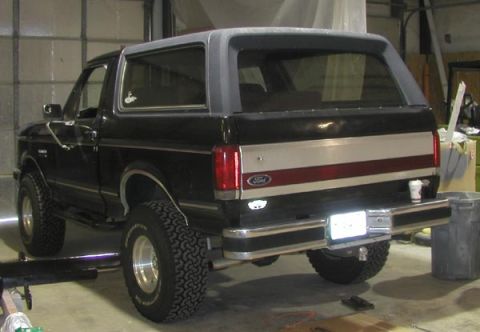 1988 Ford Bronco - Three-quarter rear view of a lifted black full-size Ford Bronco with a gray hardtop parked in a shop, sitting on chrome slotted wheels and…