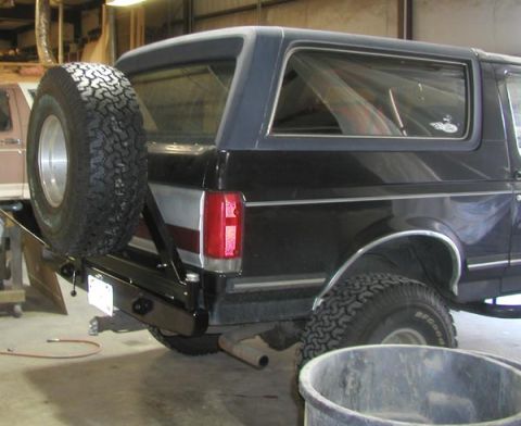 1988 Ford Bronco - Three-quarter rear view of a dark blue full-size Ford Bronco inside a shop, fitted with a fabricated black steel rear bumper and swing-out…