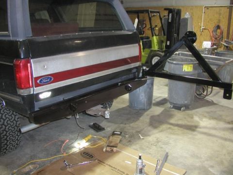 1988 Ford Bronco - Rear view of a dark full-size Ford Bronco in a shop with a new black steel rear bumper and a tubular swing-out spare carrier swung fully op…