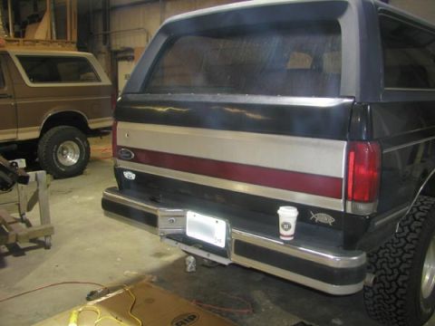 1988 Ford Bronco - Rear view of a dark full-size Ford Bronco inside a shop, still wearing its stock chrome rear bumper with a blank white license plate and a…