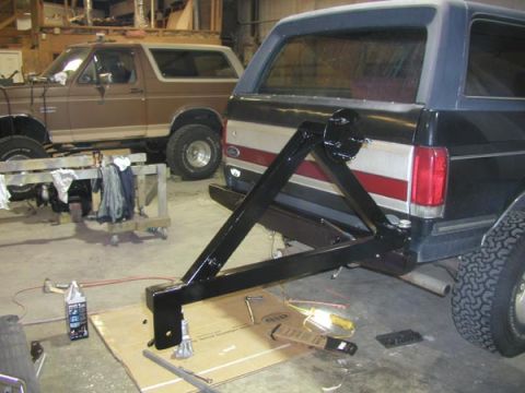 1988 Ford Bronco - Freshly painted black tubular swing-out spare tire carrier mounted to the rear bumper of a dark blue full-size Ford Bronco inside a shop, s…