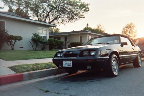 1985 Ford Mustang - Black Fox-body Ford Mustang GT convertible with the top up, parked along a residential curb at golden hour, seen from a low driver's-side f…