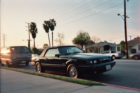 1985 Ford Mustang - Black Fox-body Ford Mustang GT convertible with tan soft top parked at a suburban curb at sunset, shot from the front three-quarter passeng…