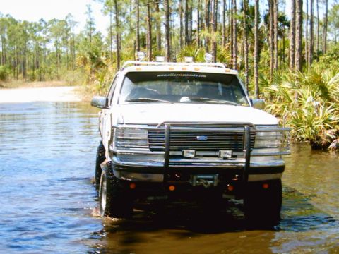 1995 Ford Bronco - Head-on view of a lifted white Ford Bronco standing in knee-deep water on a flooded sand trail through pine and palmetto woods. Visible det…