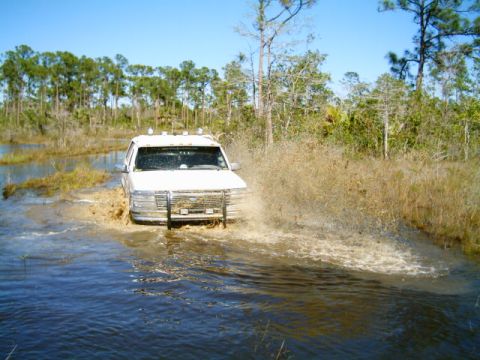 1995 Ford Bronco - White Ford truck with a tube grille guard and roof-mounted cab lights blasting through a flooded trail, throwing a large sheet of muddy spr…