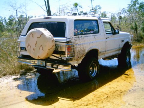1995 Ford Bronco - Rear 3/4 view of a lifted white Ford Bronco standing in shallow water on a sandy trail, mud splattered up the rocker panels and rear quarte…