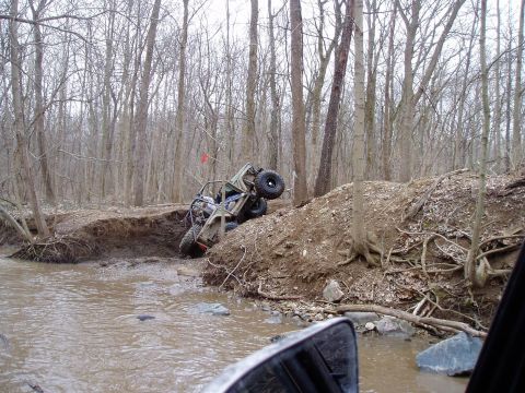 1992 Ford Bronco - An off-road vehicle is stuck in a muddy creek bed, tilted precariously on a dirt embankment surrounded by trees.