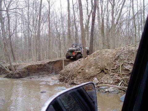 1992 Ford Bronco - An off-road vehicle navigates a muddy trail through a wooded area, with a Supermotors.net logo in the corner.