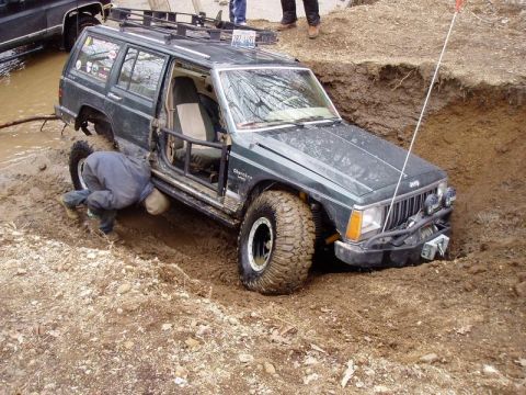 1992 Ford Bronco - A person works on a stuck Jeep Cherokee in a deep mud pit, with another vehicle nearby.