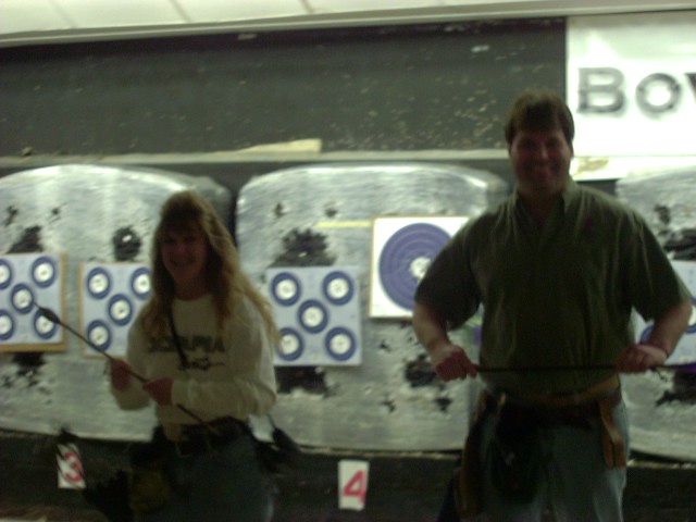 Two people stand in front of archery targets, one holding a bow