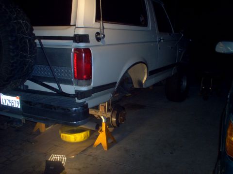 1988 Ford Bronco - The rear axle of a white Ford Bronco is raised on jack stands, with a yellow oil drain pan underneath.