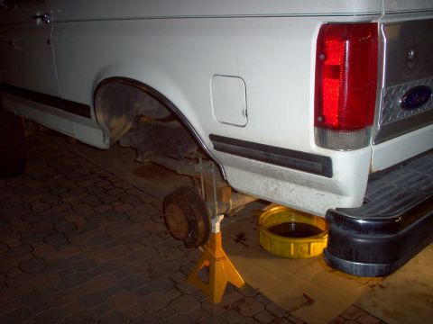 1988 Ford Bronco - Rear axle of a white Ford Bronco, lifted on jack stands for maintenance, with a yellow oil pan catching fluid.
