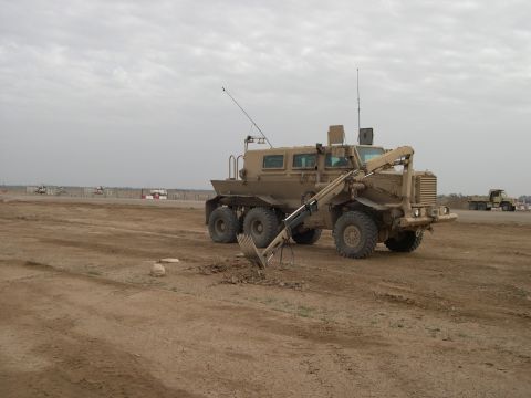 1989 Ford Bronco - A tan, six-wheeled military vehicle equipped with an excavator arm is shown in a dusty, open area with other vehicles in the distance.