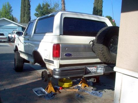 1988 Ford Bronco - A white 1988 Ford Bronco is parked on jack stands in a driveway, with its rear wheel removed for maintenance.