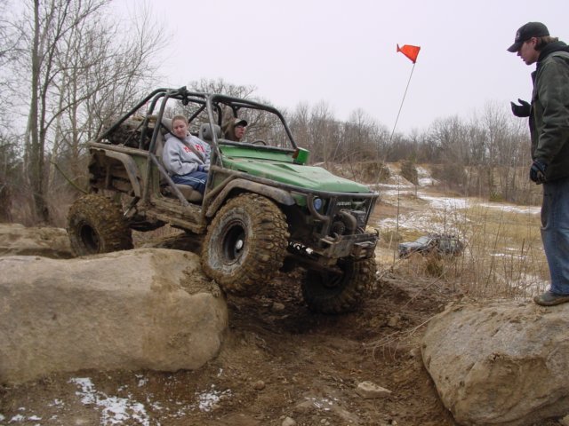 Green off-road buggy with roll cage climbing over large rocks, two people inside