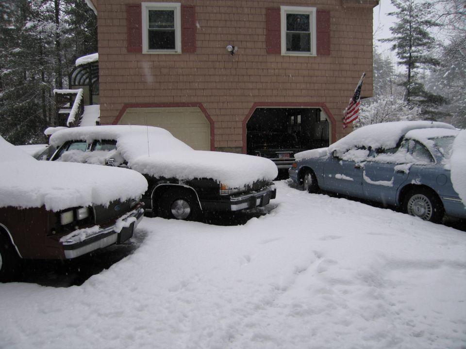 Three cars covered in snow in a driveway, with a house and garage in the background