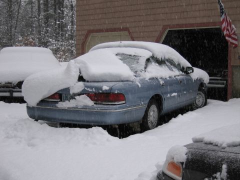 2001 Ford Crown Victoria - A blue sedan is partially buried in snow, parked in front of a garage with an American flag visible.