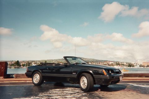 1985 Ford Mustang - A black 1985 Ford Mustang convertible is seen from the front-three-quarter view with a coastal city in the background.