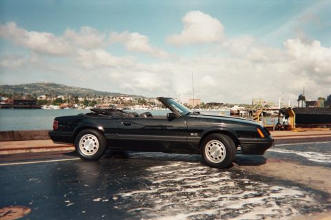 1985 Ford Mustang - Side view of a black 1985 Ford Mustang convertible with the top down, parked near a harbor.