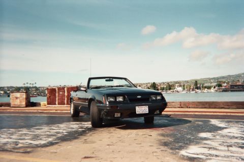 1985 Ford Mustang - A black convertible sits by the water with boats in the background under a partly cloudy sky.