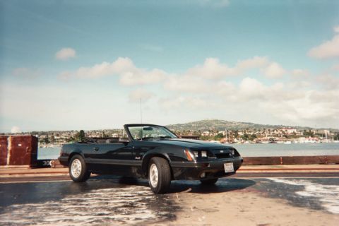 1985 Ford Mustang - A black 1985 Ford Mustang convertible is seen parked near a waterfront with a town in the background.