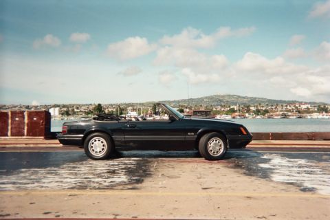 1985 Ford Mustang - A black 1985 Ford Mustang convertible is parked near a body of water with a town and hills in the distance.