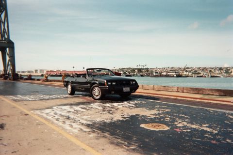 1985 Ford Mustang - A black 1985 Ford Mustang convertible is parked near a harbor with a city skyline in the background.
