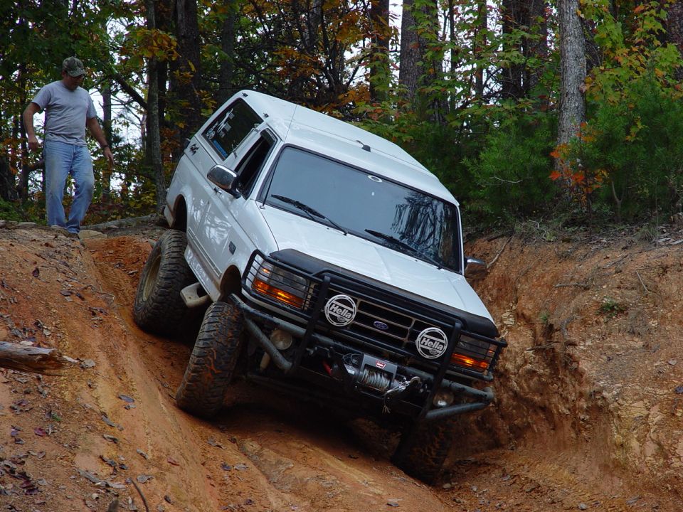 Man watches a white Ford truck climb out of a red-clay rut with brush guard and winch