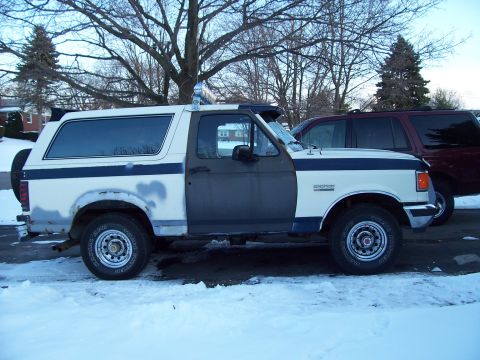 1990 Ford Bronco - A two-tone Ford Bronco sits in a snowy area, with a dark SUV parked behind it.