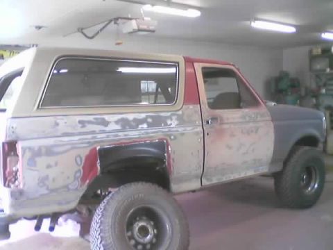 1989 Ford Bronco - Passenger-side view of a two-door full-size Ford Bronco inside a garage mid-bodywork, with red paint sanded down to primer and bare metal a…