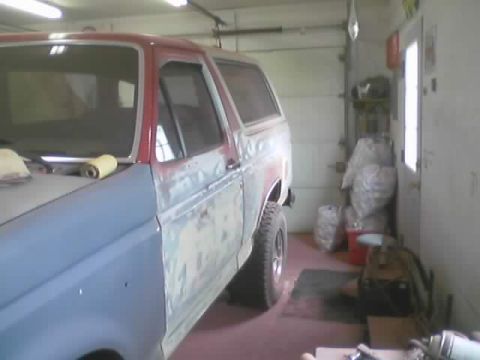 1989 Ford Bronco - Looking rearward along the driver's side of a two-door Ford Bronco in a garage during bodywork, with the red paint sanded through to gray p…