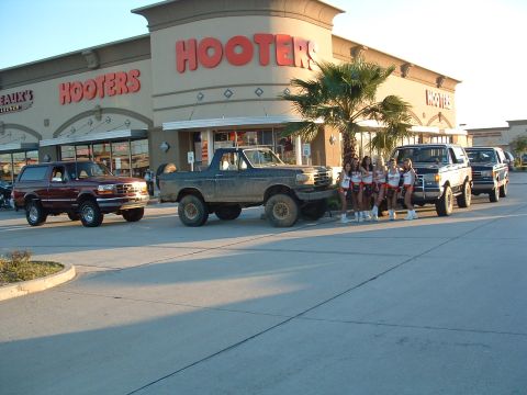 1993 Ford Bronco - A line of Ford Broncos are parked outside a Hooters, with several women posing in front of one.