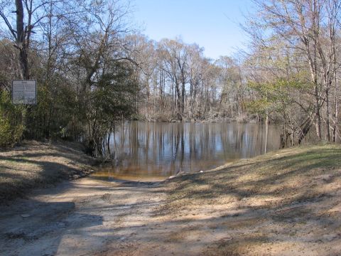 1995 Ford Bronco - A dirt boat ramp descends towards a tranquil body of water, bordered by trees under a clear sky.
